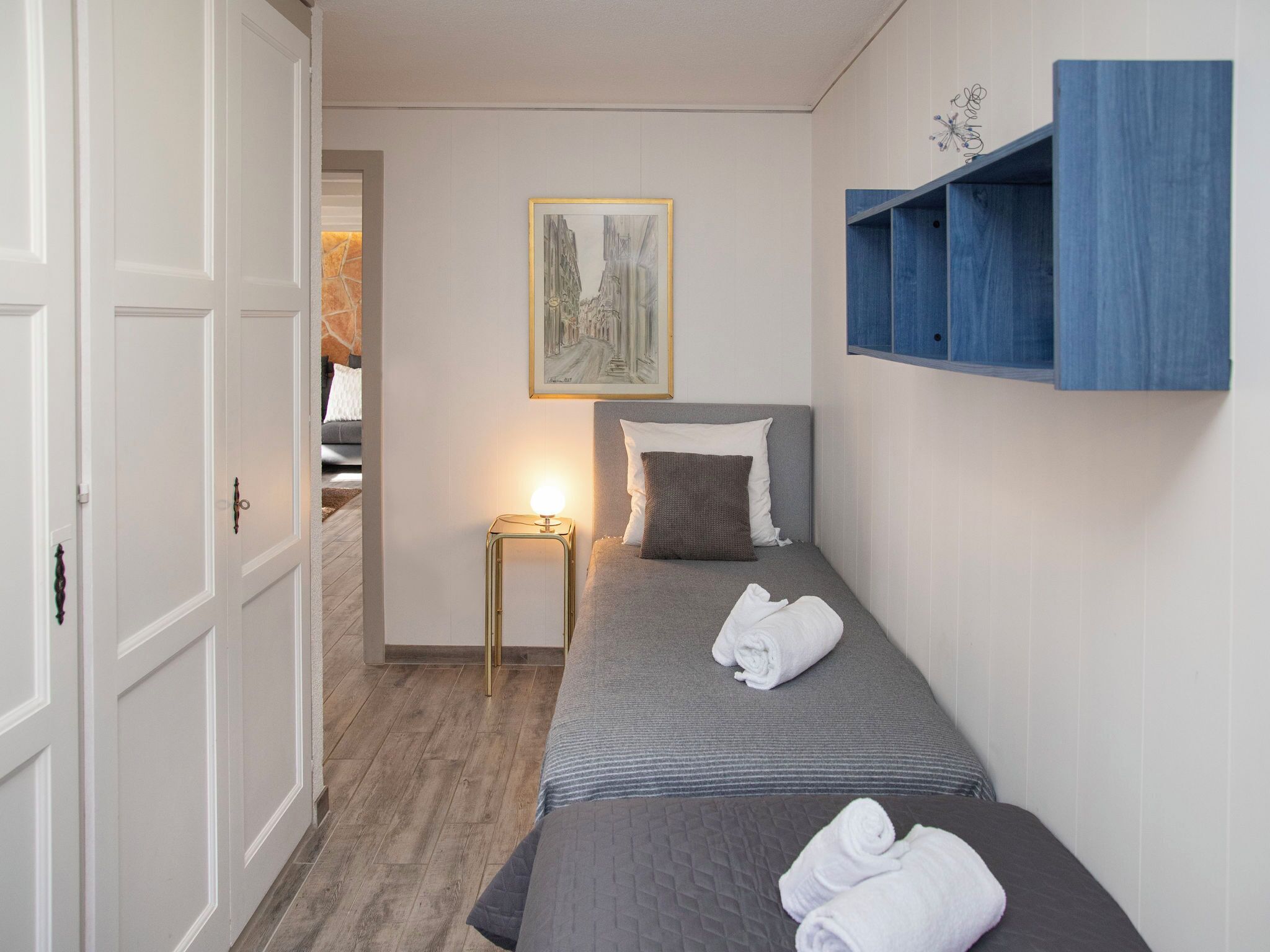 Un dormitorio con dos camas, una mesita de noche con lámpara, un cuadro en la pared y un estante azul.