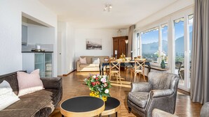 A spacious living area with a sofa, two round tables, and a dining table with chairs. There is a kitchen area with a cabinet and a window with a view of mountains. - Suite Modern by Interhome (Ascona)