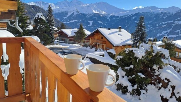 Eine Holzdar Terrasse mit zwei weißen Tassen, mit Blick auf schneebedeckte Häuser und Berge.