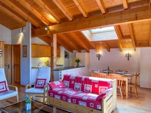 Een gezellige kamer met een houten plafond, een bank met een gestreept hoesje, een eettafel met stoelen, en een keukenhoek met kastjes en een spoelbak.