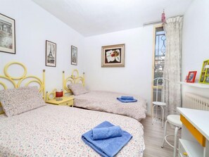 Slaapkamer met twee bedden, een bureau en een stoel. Er hangen afbeeldingen in lijsten aan de muur en een raam met gordijnen.