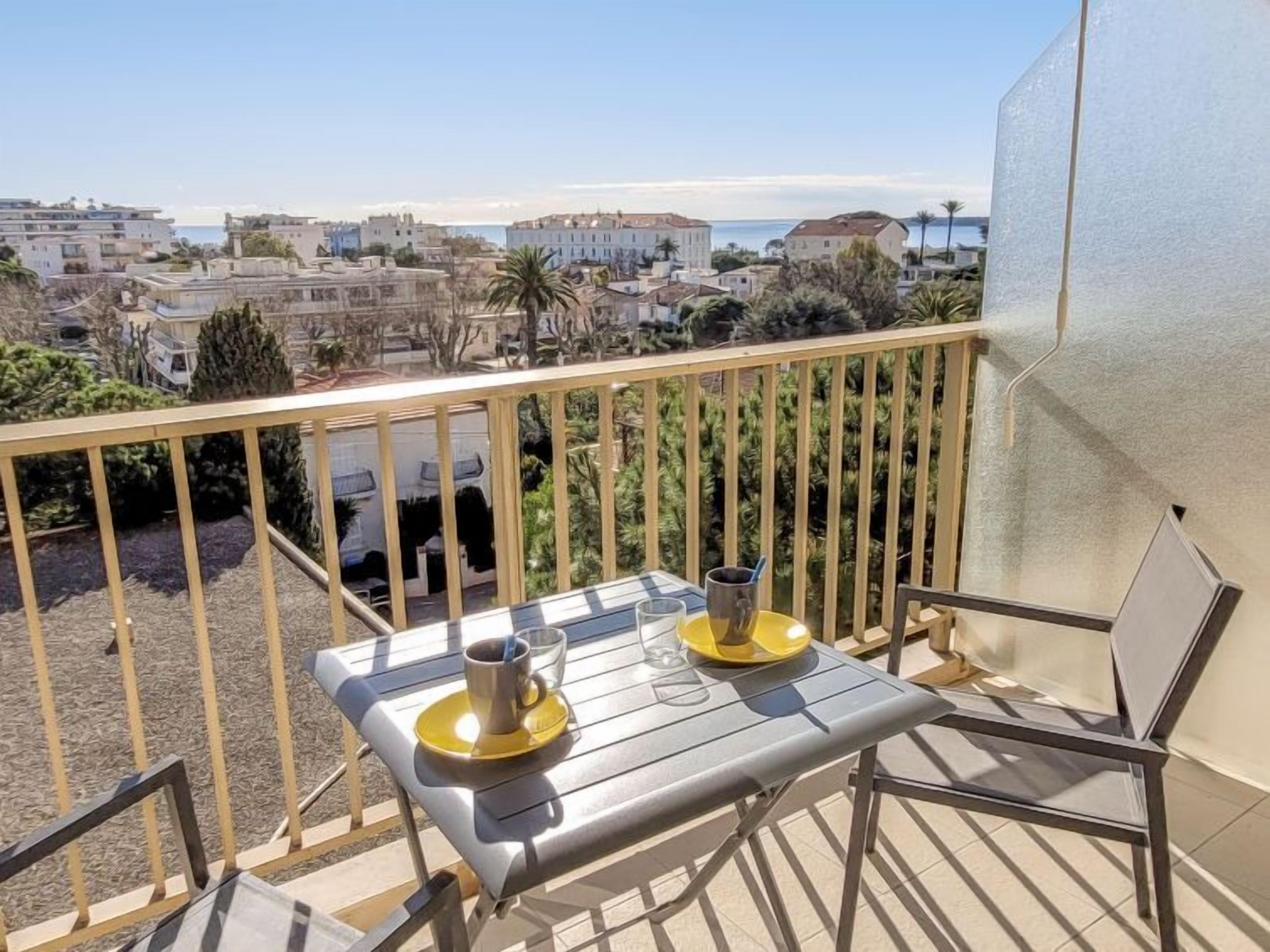 A balcony with a table and chairs, overlooking a coastal town with buildings and a clear sky.