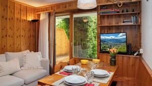 Une pièce chaleureuse avec un décor intérieur en bois, une table à manger dressée pour deux personnes, un canapé et un téléviseur diffusant un paysage pittoresque.