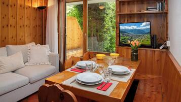 Une pièce chaleureuse avec un décor intérieur en bois, une table à manger dressée pour deux personnes, un canapé et un téléviseur diffusant un paysage pittoresque.