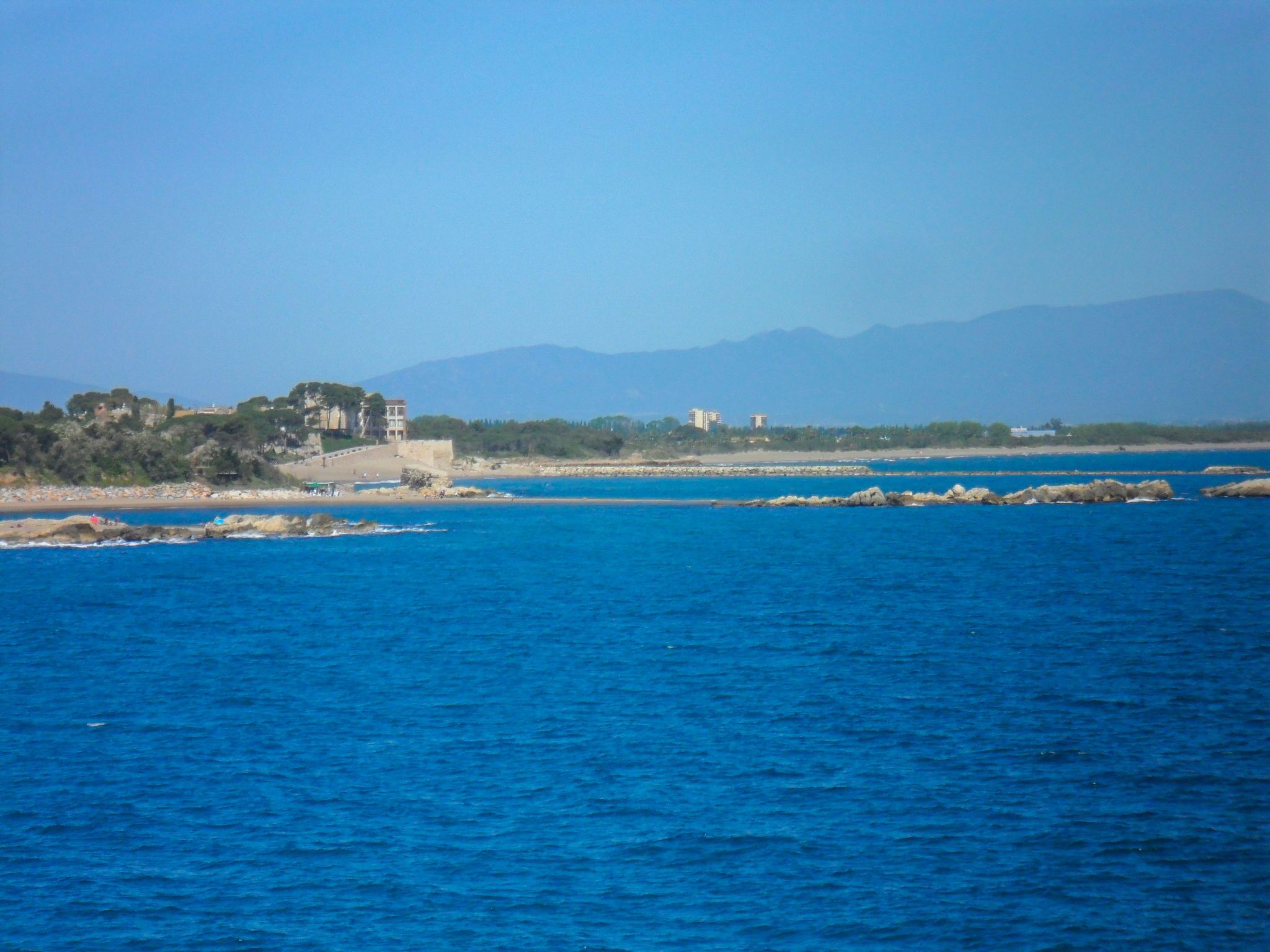 A coastal view with clear blue waters, rocky outcrops, and buildings along the shore.