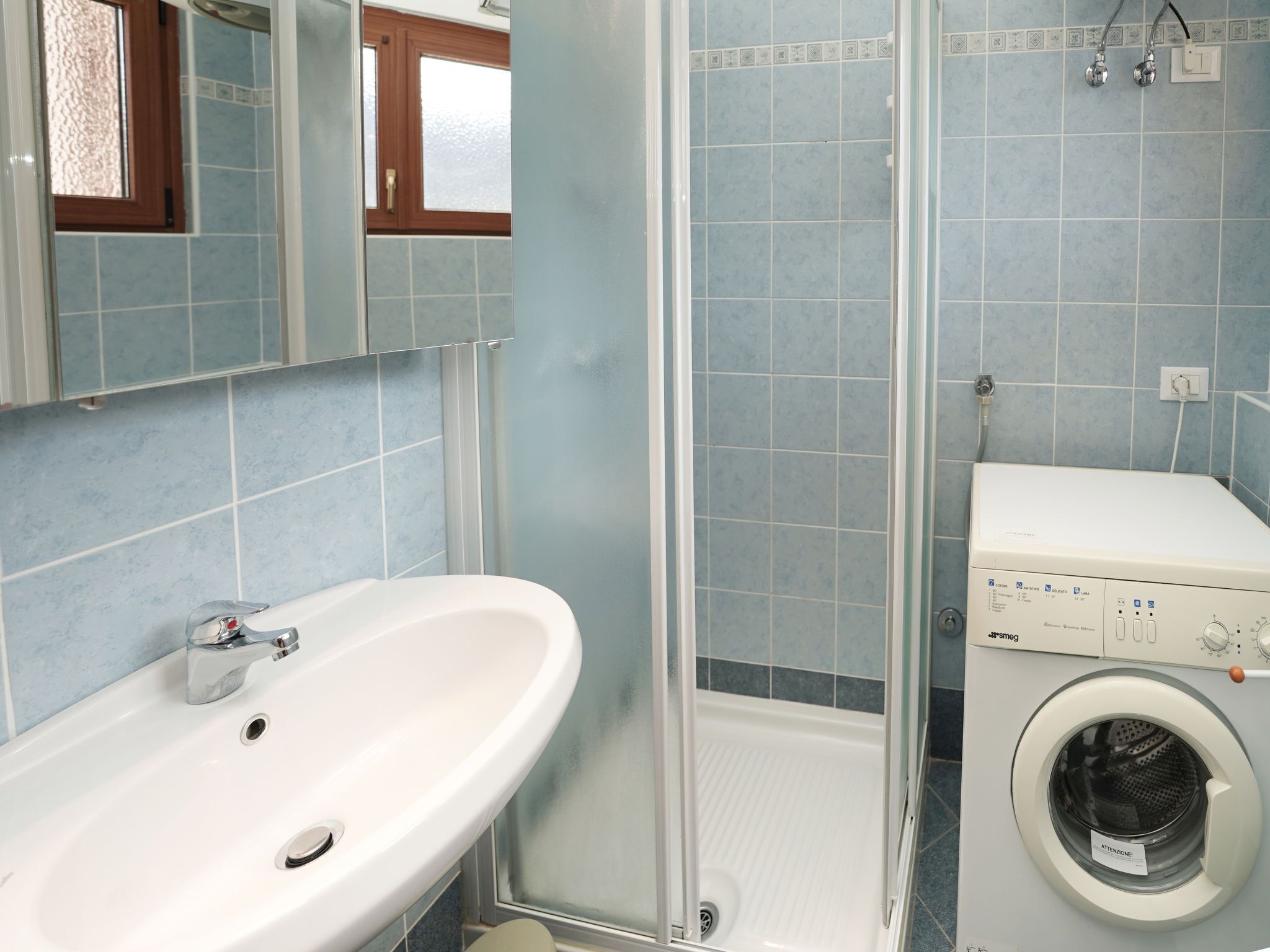 Un baño con un lavabo blanco, cabina de ducha con mampara de vidrio y lavadora de carga frontal.