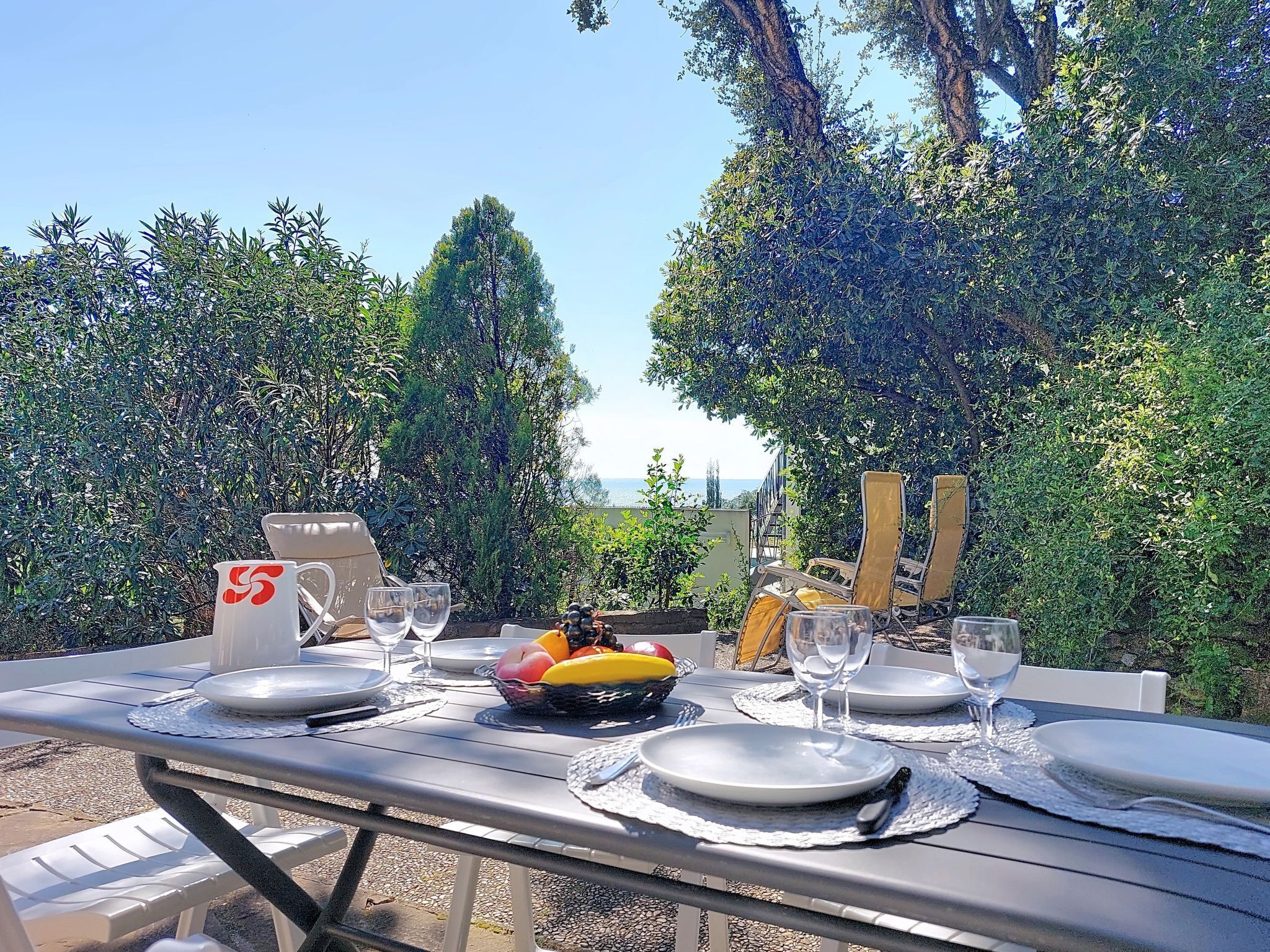 An outdoor dining table set with plates, glasses, and a fruit basket, surrounded by lush greenery and a view of the sea.