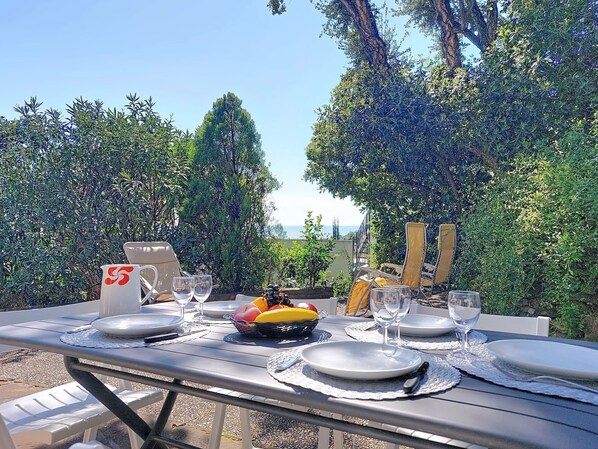 An outdoor dining table set with plates, glasses, and a fruit basket, surrounded by lush greenery and a view of the sea.