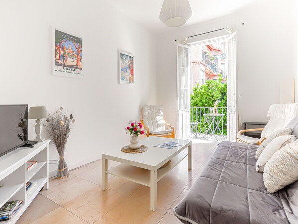 A bright living room with a sofa, a TV, a table with a vase of flowers, and a balcony with a view of greenery. - Apartment Hérold by Interhome (Nice)