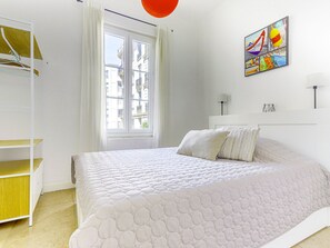 A bedroom with a bed, a window with curtains, a wall-mounted artwork, and a shelf with clothes. - Apartment Hérold by Interhome (Nice)
