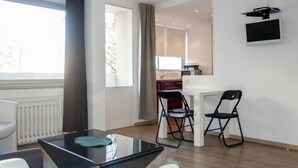 Una camera moderna con un tavolino da caffè in vetro, un divano bianco e un'area pranzo con due sedie. Si intravede una cucina sullo sfondo.