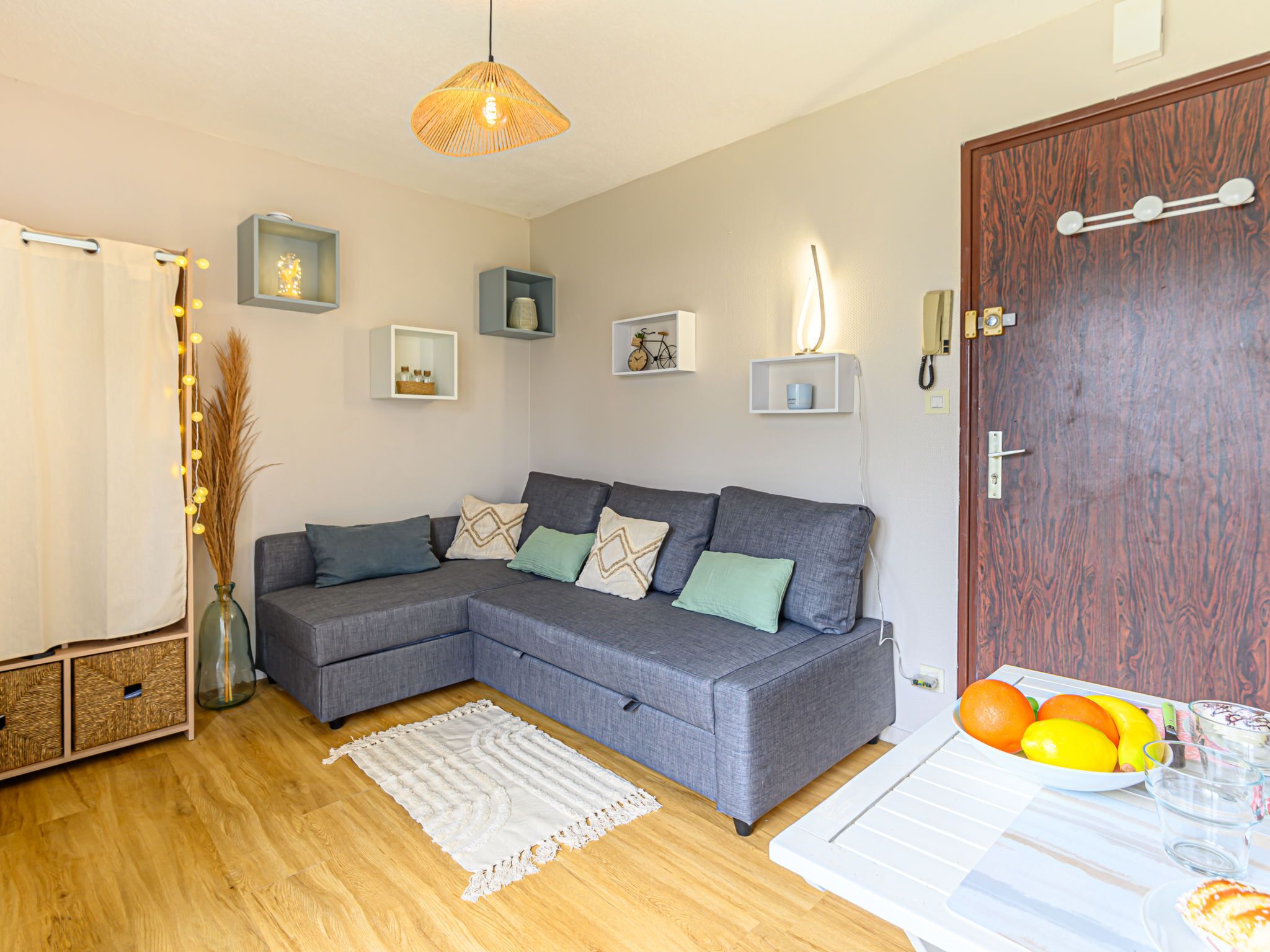 Un salon avec un canapé d’angle gris, un tapis tissé et un plancher en bois. On y trouve des étagères murales ornées d’objets décoratifs et une porte avec un interrupteur.
