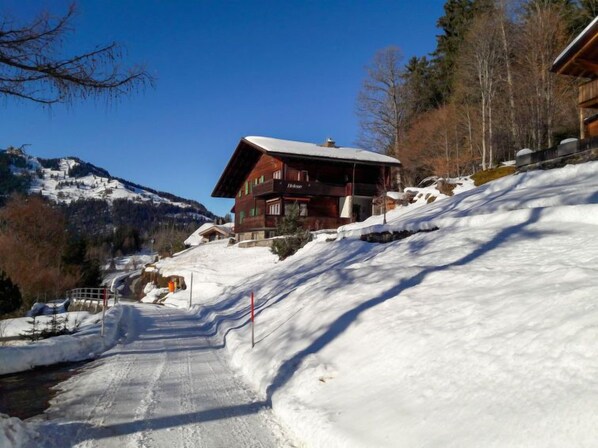 A wooden chalet with a sloped roof, situated on a snow-covered hillside with a mountain in the background.