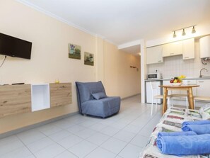 Un espace de vie compact comprenant un canapé, une table à manger avec un panier de fruits, un coin cuisine et un téléviseur à écran plat fixé au mur.