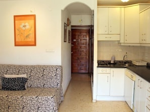 A compact living space with a patterned sofa, a kitchen with white cabinets, and a framed floral artwork on the wall.