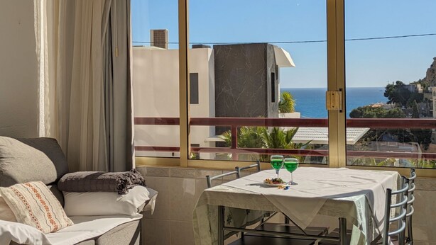 Ein Balkon mit Tisch für zwei Personen, einer Couch und Blick auf das Meer und Gebäude.