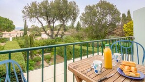 Un balcón con mesa lista para el desayuno, con vista a un jardín con árboles y una vista distante de edificios.
