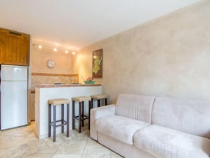 A modern kitchen with a white refrigerator, wooden cabinets, and a tiled backsplash. There are three bar stools and a white sofa in the living area.