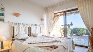 A bedroom with a large bed, a nightstand, a lamp, and a view of the outdoors.