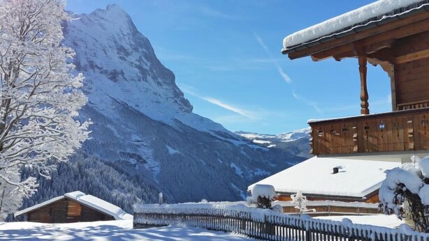 A wooden chalet with a snow-covered roof, surrounded by a wooden fence and a snow-covered mountain in the background. - Lohnerhus by Interhome (Grindelwald)