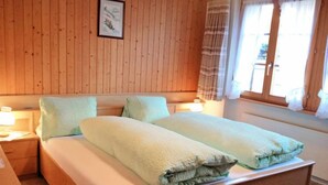 A double bed with a light-colored duvet and pillows, wooden headboard, bedside tables with lamps, and a window with curtains. - Lohnerhus by Interhome (Grindelwald)