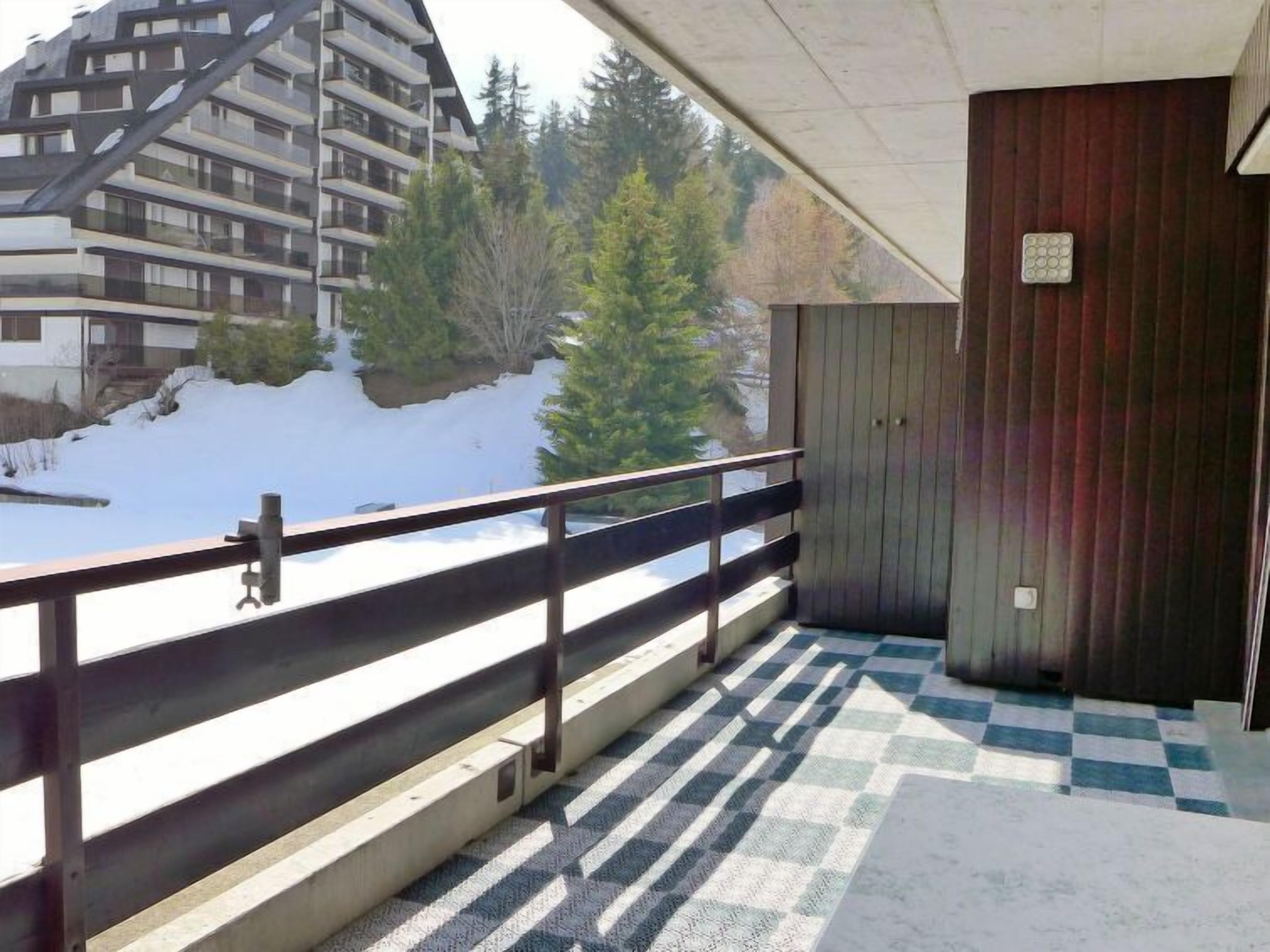 A balcony with a checkered floor, a wooden railing, and a view of a building and trees in the background.