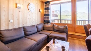 A wooden interior with a leather sofa, a table with cups, a clock, and a window with a view of a building and landscape. - Lac Blanc 704 by Interhome (Val Thorens)