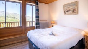 A bedroom with a large bed, a window with a view of mountains, a painting on the wall, and a wooden ceiling. - Lac Blanc 704 by Interhome (Val Thorens)