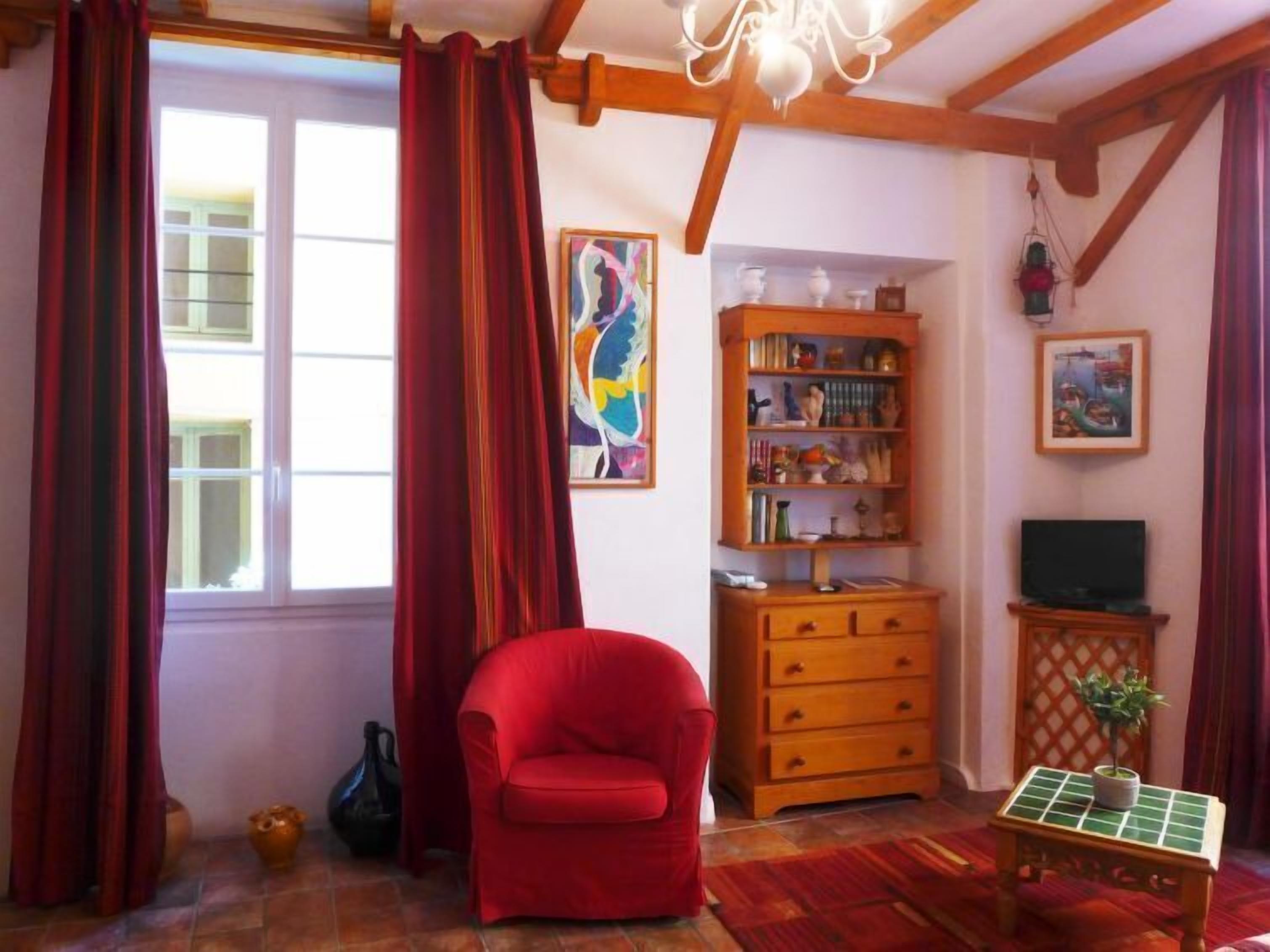 A cozy living room with a red armchair, wooden bookshelves, a television, and a window with red curtains.