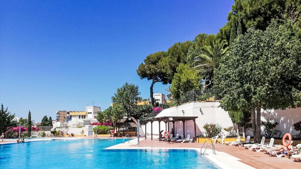 Una piscina con lettini e ombrelloni, circondata da alberi ed edifici.
