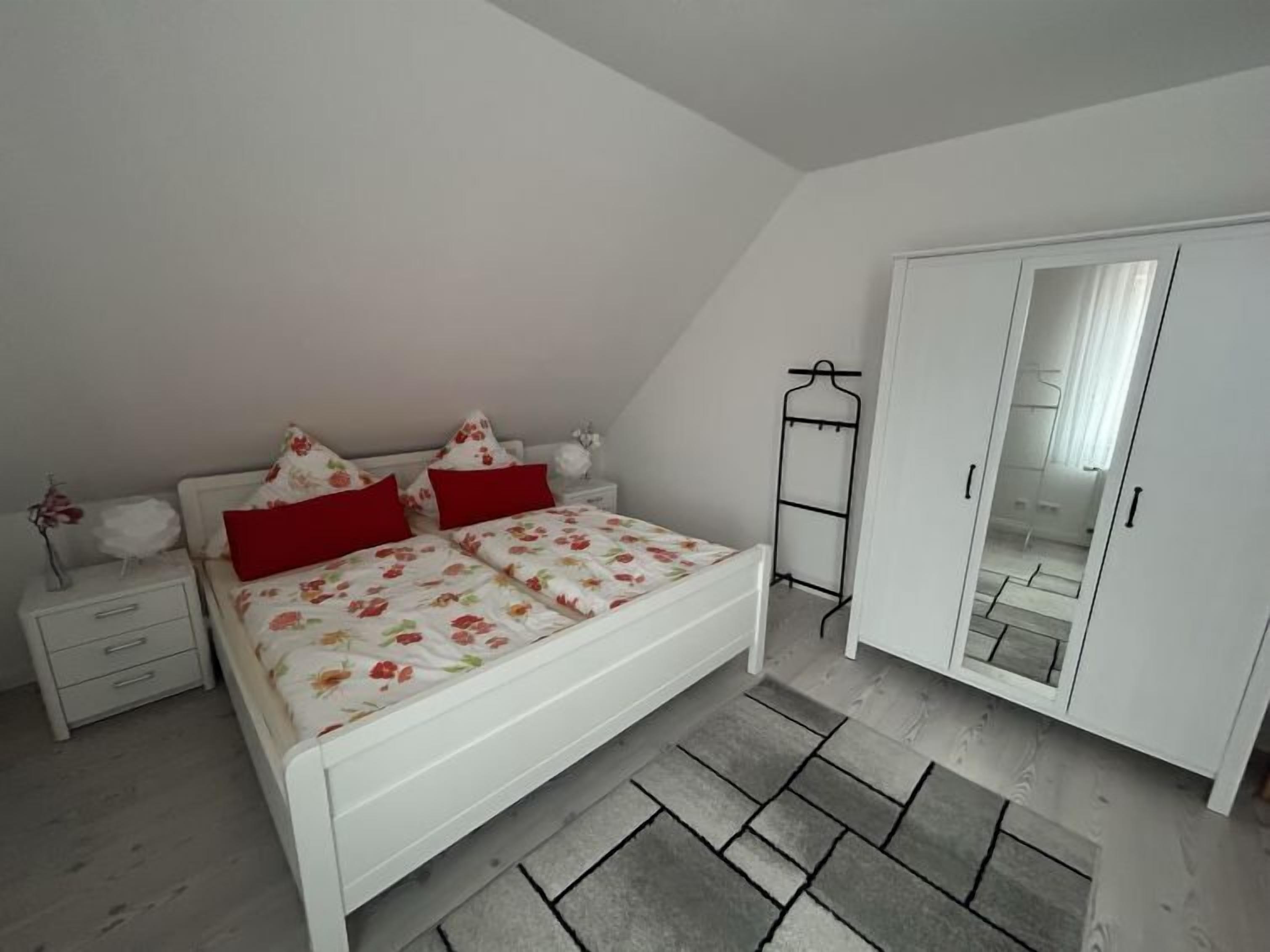 A bedroom with a white bed, red pillows, a white wardrobe, and a mirror.