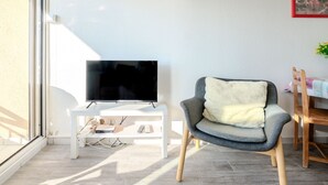A modern living room with a flat-screen TV on a white stand, a grey armchair with a fluffy pillow, and a dining table with a wooden chair. - les Goelands by Interhome (Mimizan)
