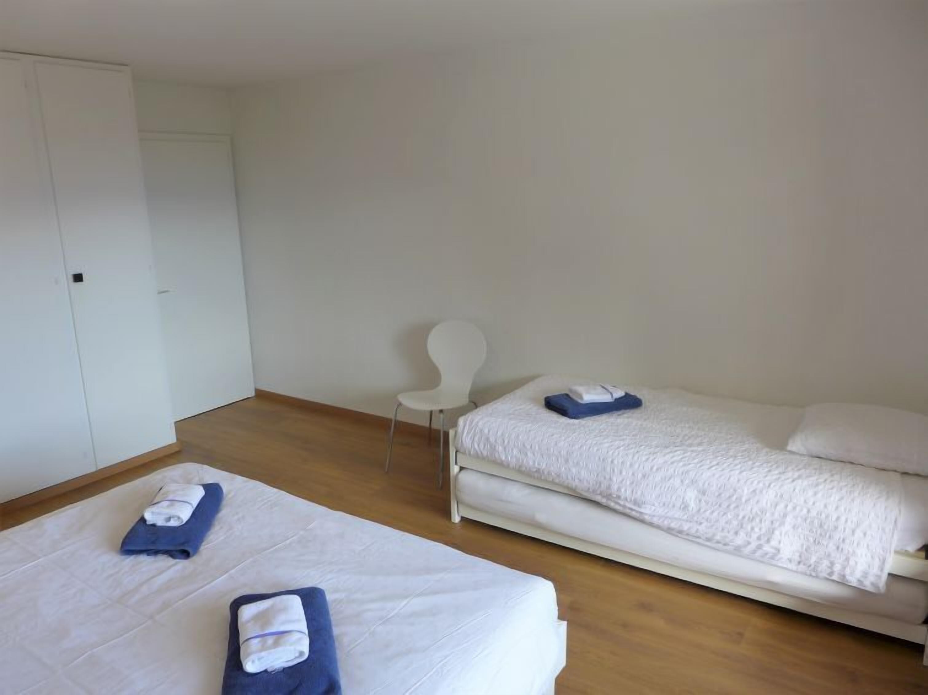 A hotel room with two beds, a chair, and a wardrobe.