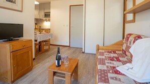 A compact room with a bed, a TV, a small table with a bottle and glasses, a wooden dresser, and a kitchenette in the background. - Balcons d'Olympie 305 - A by Interhome (Les Menuires)