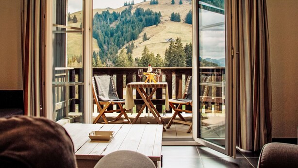 A room with a wooden table, two chairs, and a view of a mountainous landscape through large windows. - Andromède 3-pièces A34 by Interhome (Moléson-sur-Gruyères)