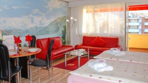 A hotel room with a bed, a red sofa, a dining table with chairs, a painting of a mountain landscape, and a window with curtains.