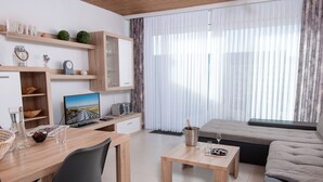 Uma sala de estar moderna com uma escrivaninha de madeira, uma cadeira preta, um sofá cinzento e uma mesinha de centro. Há uma televisão na escrivaninha, uma janela com cortinas leves e uma garrafa de champanhe na mesinha de centro.