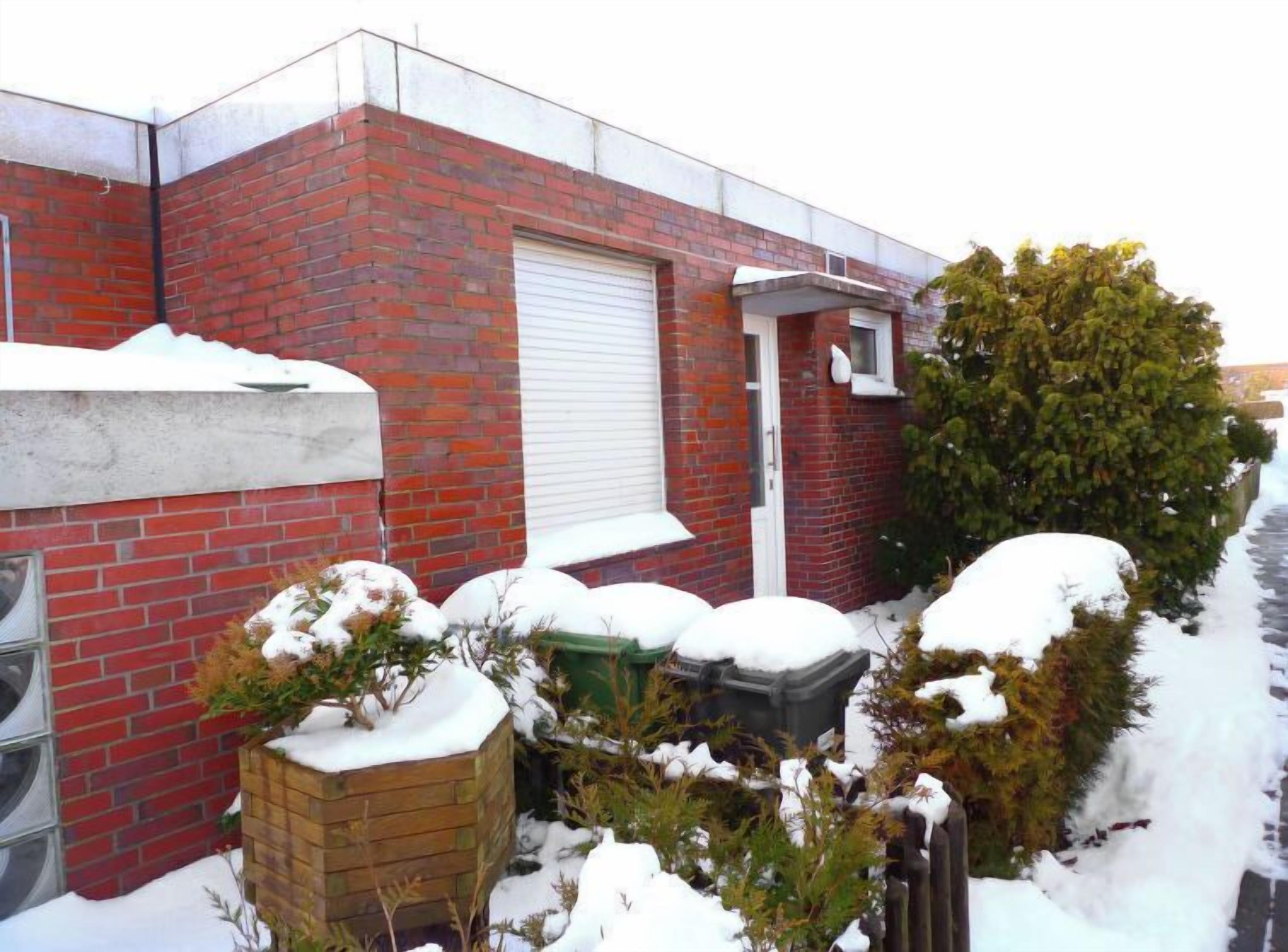 Un edificio de ladrillo con una puerta de garaje blanca, una ventana pequeña y un jardín cubierto de nieve con plantas y una jardinera de madera.
