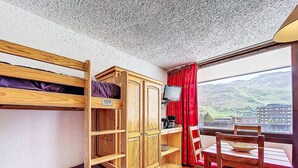 A room with bunk beds, a wooden cabinet, a table with chairs, and a view of a mountainous landscape. - Aravis 101 by Interhome (Les Menuires)