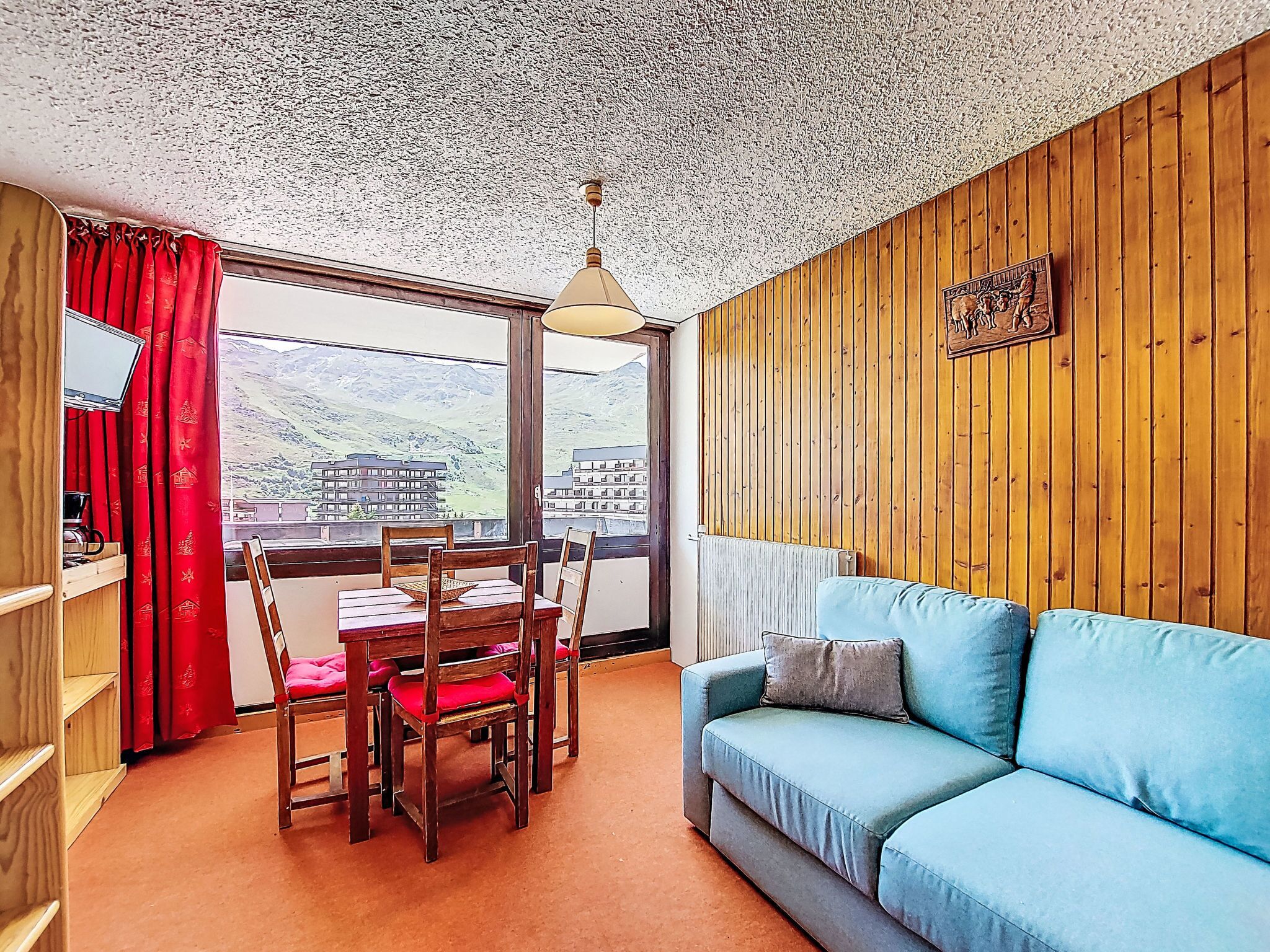 A cozy room with a wooden wall, a blue sofa, a dining table with chairs, and a view of a mountain landscape through a large window.