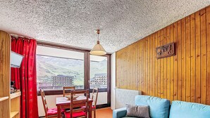 A cozy room with a wooden wall, a blue sofa, a dining table with chairs, and a view of a mountain landscape through a large window. - Aravis 101 by Interhome (Les Menuires)