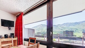 A balcony with a wooden table and chairs, a television, and a view of a mountainous landscape. - Aravis 101 by Interhome (Les Menuires)