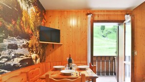 A wooden dining area with a table set for two, a television mounted on the wall, and a view of a grassy landscape through an open door. - Penia 2 by Interhome (Canazei)
