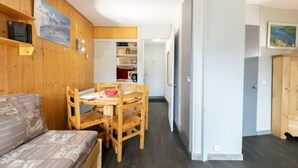 Ett kompakt rum med trägolv, en säng, ett matbord med stolar, en köksdel och en skjutdörr som leder till ett annat rum.