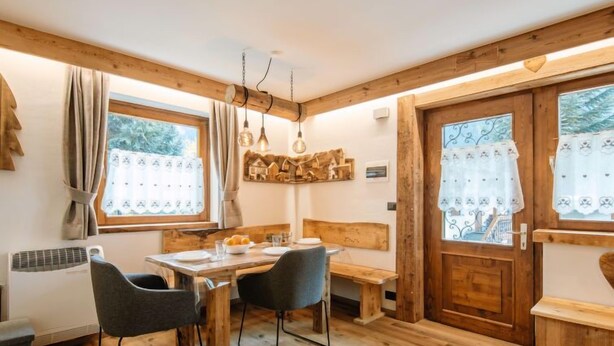 Un coin repas chaleureux, avec des meubles en bois, une table dressée pour deux personnes et un banc. Il y a deux fenêtres avec des rideaux, dont l’une offre une vue sur la verdure à l’extérieur.