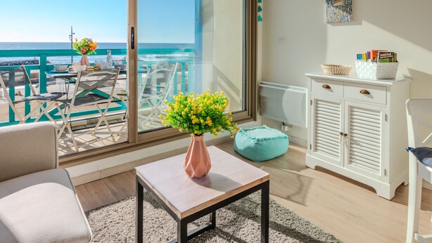 Un balcon avec vue sur la mer, une table ornée d’un vase de fleurs, une armoire blanche remplie de livres et un pouf turquoise.