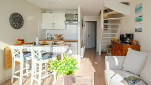 Une cuisine moderne avec des meubles blancs, un coin repas avec des chaises blanches, une machine à café et un escalier menant à une chambre en mezzanine.