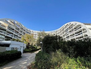 A modern, curved building with multiple balconies, surrounded by greenery and a clear sky.
