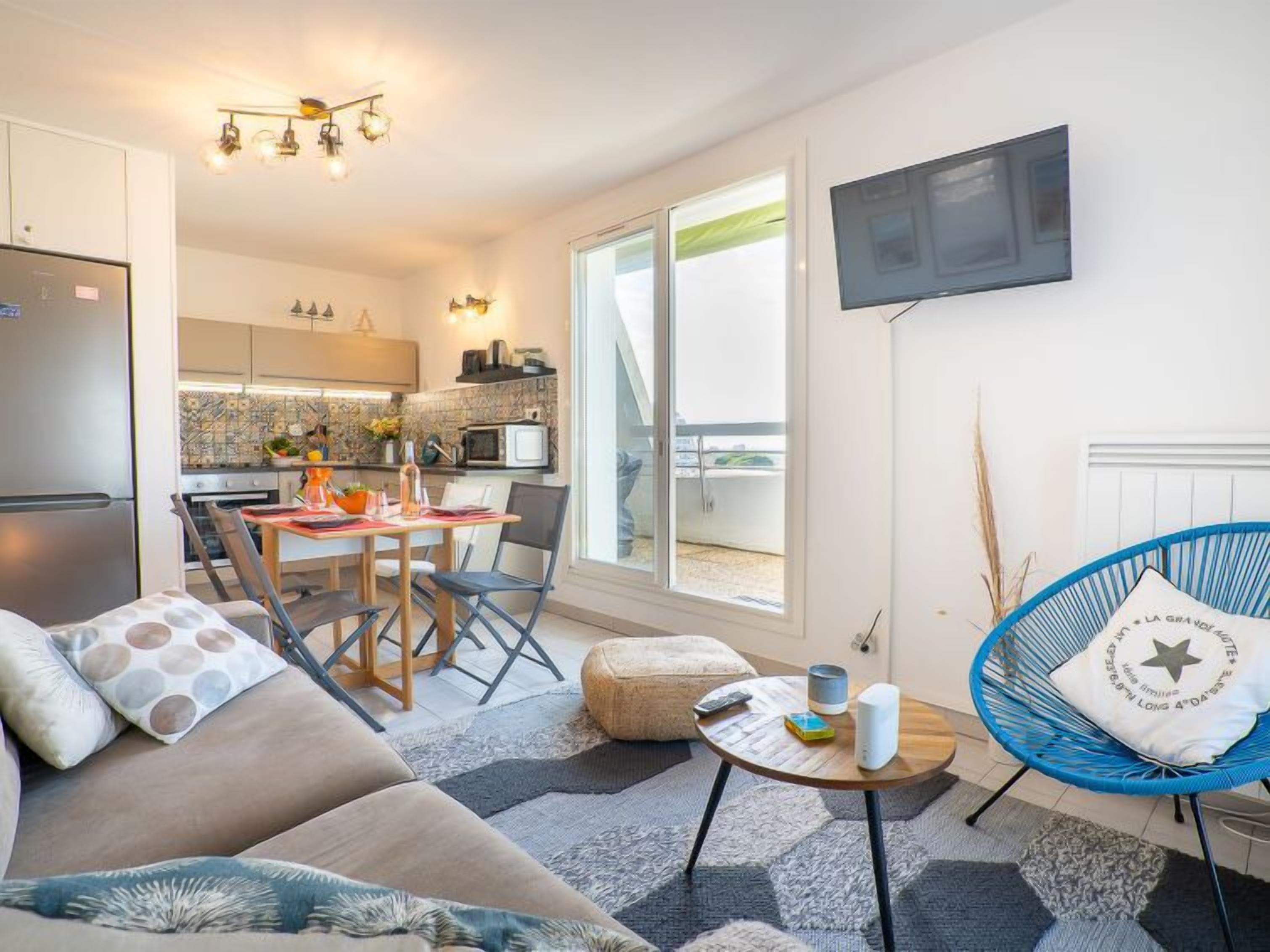 A modern living room with a sofa, a round coffee table, and a blue chair. There is a kitchen in the background with a dining area.