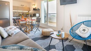 Ein modernes Wohnzimmer mit einer Couch, einem runden Couchtisch und einem blauen Sessel. Im Hintergrund ist eine Küche mit Essbereich zu sehen.